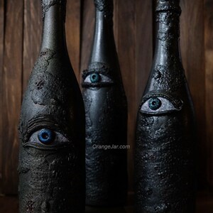 May include: Three black glass bottles with blue eyes painted on them. The bottles are decorated with a textured, rough surface. The bottles are arranged in a row on a wooden surface. The text "OrangeJar.com" is visible on the surface.