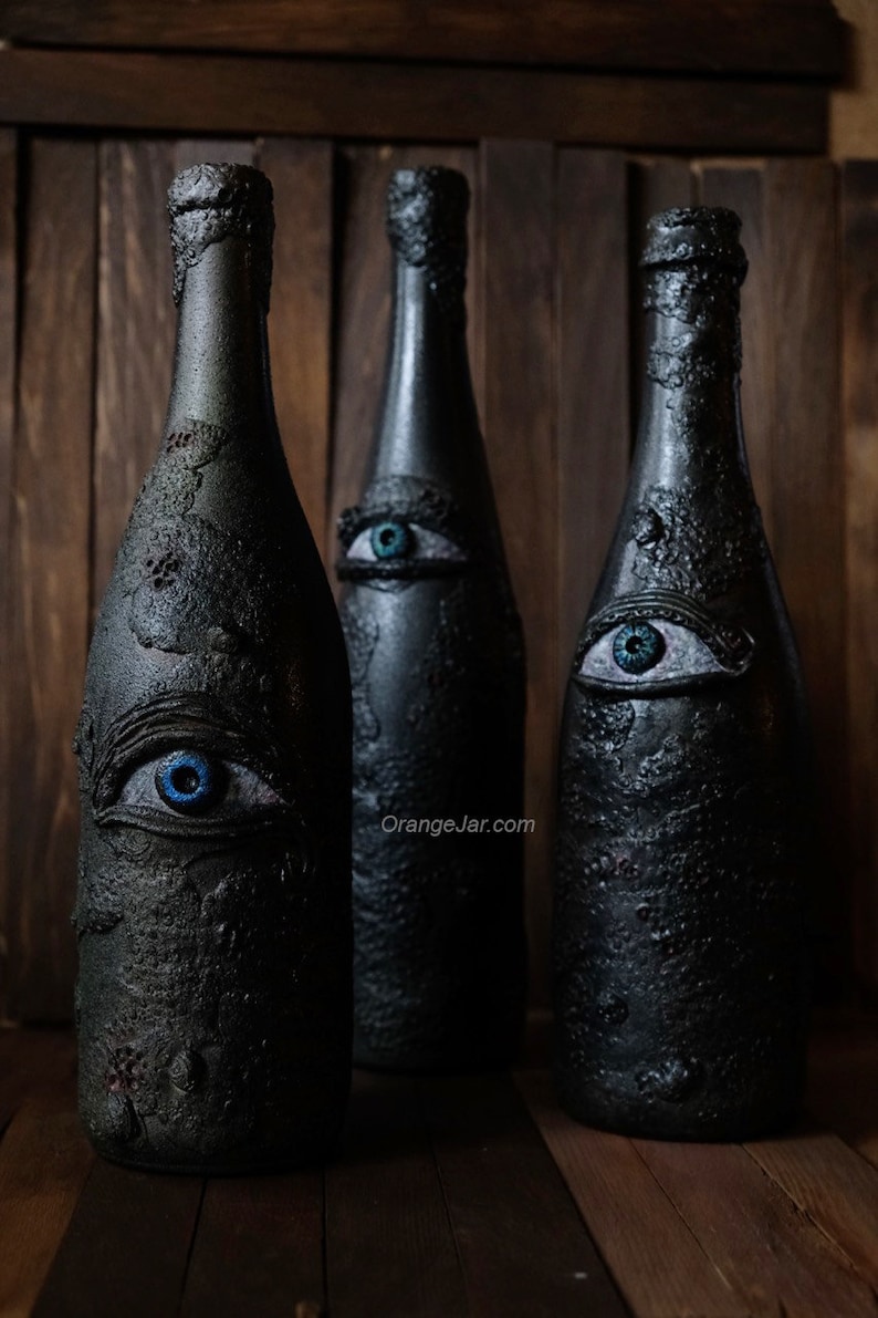 May include: Three black glass bottles with blue eyes painted on them. The bottles are decorated with a textured, rough surface. The bottles are arranged in a row on a wooden surface. The text "OrangeJar.com" is visible on the surface.