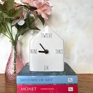 May include: A white ceramic house-shaped clock with black hands and the numbers twelve, nine, three, and six. The clock sits atop two books, one blue and one red, with a pink vase and flowers in the background.