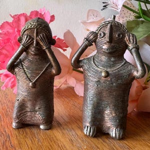 May include: Two bronze-toned figurines, one covering its eyes and the other its ears. Both have detailed, textured bodies and wear necklaces. The figures are posed in a 'see no evil, hear no evil' stance, with a decorative floral background.