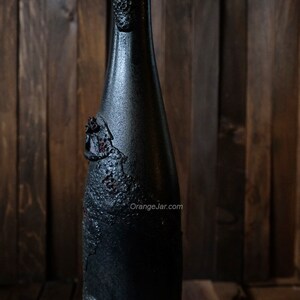 May include: A black glass bottle with a textured surface and a dark brown, almost black, color. The bottle has a unique, almost cracked, design with a rough texture. The bottle is standing on a wooden surface.