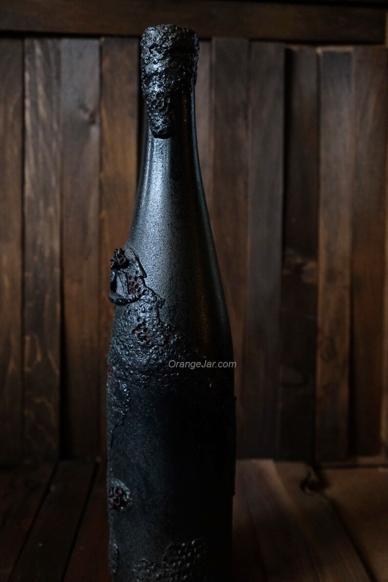 May include: A black glass bottle with a textured surface and a dark brown, almost black, color. The bottle has a unique, almost cracked, design with a rough texture. The bottle is standing on a wooden surface.