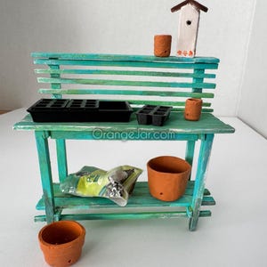 May include: A miniature wooden potting bench with a teal green finish. The bench has two shelves and a back with slatted wood. There are several small terracotta pots, a birdhouse, and seed packets on the bench.