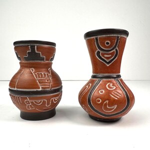 May include: Two terracotta vases with dark brown rims and bases, featuring intricate white etched geometric and figurative patterns. The shorter vase has a wide body, while the taller vase has a slender neck and a rounded base, both displaying detailed carvings.