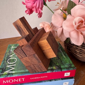 May include: A wooden puzzle sits atop two books, "MONET" and "HISTORY OF ART." The puzzle is constructed of interlocking wooden blocks in shades of brown. Pink flowers and a wicker basket are in the background.