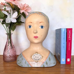 May include: A hand-painted mannequin head with blue eyes, pink lips, and a decorative headband. The bust features a blue and green dress design with the text "coiffure pour madeleine" on a small plaque. Two books stand to the right.