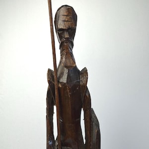 May include: A dark brown hand-carved wooden figurine depicting a tall, slender figure with a pointed beard and angular facial features. The figure holds a long, thin wooden staff in the right hand and carries a shield at the left side.