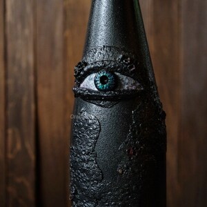 May include: A black glass bottle with a textured surface and a large, blue eye painted on the front. The eye has a black pupil and a white iris.