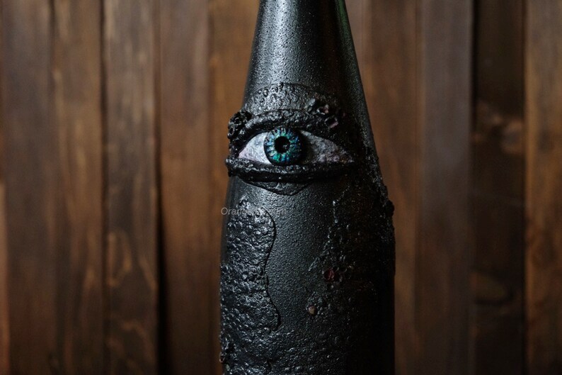 May include: A black glass bottle with a textured surface and a large, blue eye painted on the front. The eye has a black pupil and a white iris.