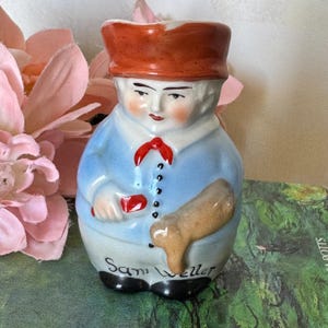 May include: A vintage ceramic figurine of a man in a blue coat, red hat, and black shoes. The figurine has a white face with rosy cheeks and a red bow tie. The name "Sam Weller" is written on the base.