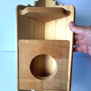 May include: A light-colored wooden corner shelf featuring a top flat surface and a lower section with a circular cutout. The unit has a vertical back panel with decorative scalloped edges at the top and bottom, designed to fit into a corner space.