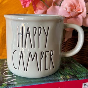 Puede incluir: Una taza de cerámica verde claro con las palabras "HAPPY CAMPER" en letras negras. La taza tiene forma redondeada y un asa. Se encuentra sobre una pila de libros, con flores rosas en el fondo.
