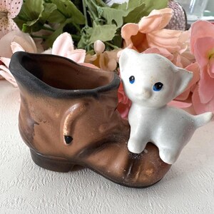 Puede incluir: Una maceta de cerámica vintage con forma de bota marrón con borde negro. Un pequeño gatito de cerámica blanca con ojos azules está posado en la bota. La maceta es para plantas o flores pequeñas.