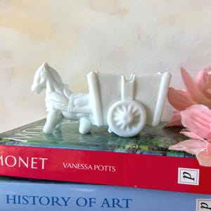Op de afbeelding: Een wit keramieken paard en wagen beeldje. Het paard is aangespannen aan de wagen, die aan de zijkant een decoratief bloemmotief heeft. De wagen heeft een spaakwiel. Het beeldje staat op een boek.