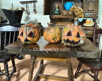 Può includere: Tre zucche arancioni intagliate in miniatura con tratti del viso neri sono su un tavolo di legno. Il tavolo è in un ambiente di cucina in miniatura, con un mobile e sedie di legno. L'indirizzo del sito web "OrangeJar.com" è stampato sul tavolo.