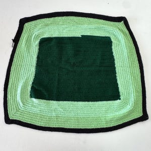 Op de afbeelding: Een handgemaakte gehaakte deken met een vierkant ontwerp. De deken heeft een donkergroen midden, omgeven door een lichtgroene rand en een zwarte rand. De deken is gemaakt van zacht garen.