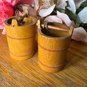 May include: Two small, wooden buckets with a light brown finish and darker brown bands. Each bucket has a small handle made of a thin wooden rod and rope. The buckets are sitting on a wooden surface.