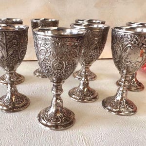 Puede incluir: Un conjunto de copas antiguas de plata con diseños florales y de volutas ornamentadas. Las copas presentan un patrón detallado en relieve y se asientan sobre bases de pedestal decorativas. El acabado plateado les da un aspecto vintage.