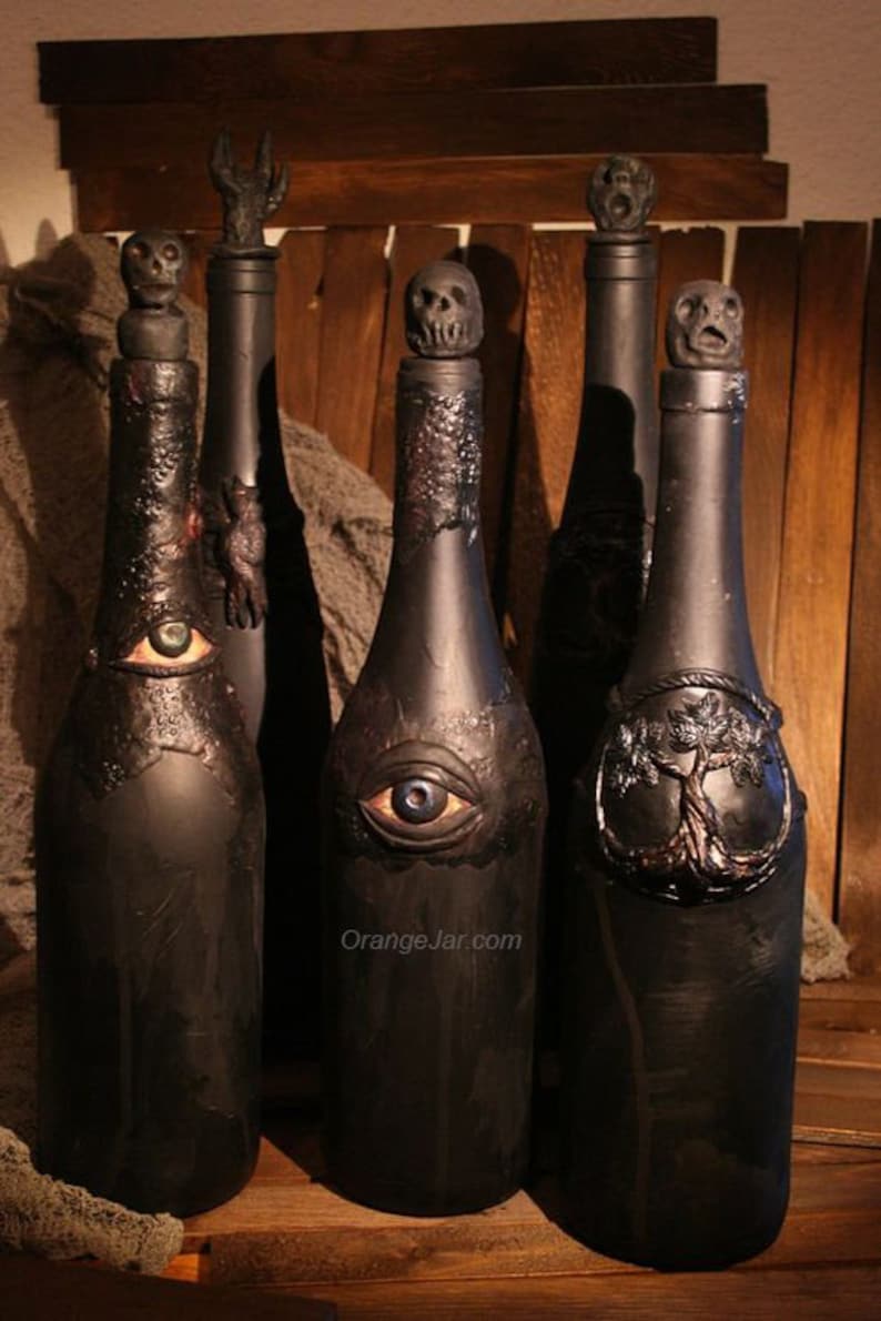 May include: Five black glass bottles with different decorative tops, including a skull, a tree, and an eye. The bottles are arranged in a row on a wooden surface. The text "OrangeJar.com" is visible on the bottom of the image.