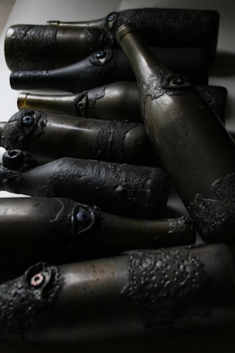 May include: A collection of dark gray glass bottles with textured surfaces and decorative eyes. The bottles are arranged in a row, with the largest bottle in the center.