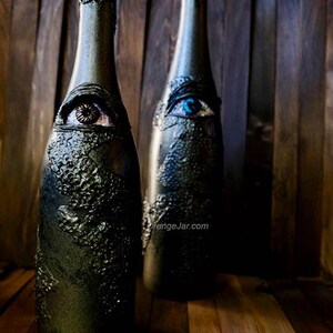 May include: Two glass bottles with a textured black finish and painted eyes. One eye is blue and the other is brown. The bottles are on a wooden surface.