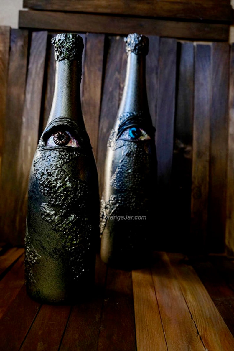 May include: Two glass bottles with a textured black finish and painted eyes. One eye is blue and the other is brown. The bottles are on a wooden surface.
