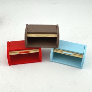 May include: Three miniature bread boxes in red, brown, and blue. Each box has a curved top and a pull-down door with a light-colored wooden accent. The boxes are made of a solid material and are designed to look like miniature bread storage containers.