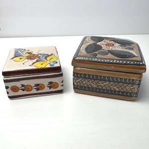 May include: Two rectangular ceramic boxes with lids. The left box is cream-colored with a yellow and blue floral design on the lid and orange circular motifs on the side. The right box is tan with a dark floral pattern on the lid and geometric lines.