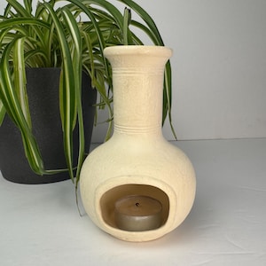 May include: A cream-colored ceramic oil burner with a textured, matte finish. The vessel features a tall, cylindrical neck with two horizontal etched lines and a rounded base with a wide, arched opening containing a small, round metal tea light.
