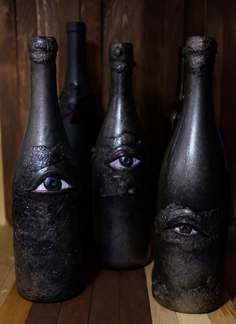 May include: Four black glass bottles with a textured surface and painted eyes. The bottles are arranged in a row on a wooden surface.