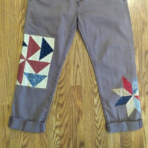 May include: A pair of gray denim jeans with patchwork fabric on the knees. The patchwork fabric is in a red, white, and blue color scheme and features a star and a pinwheel design.