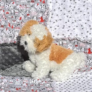 May include: A plush toy dog with white and brown fur sits on a patchwork quilt. The quilt features various dog-themed patterns in gray, white, and red, including dog illustrations, stars, and polka dots. The toy dog is a soft, cuddly item.