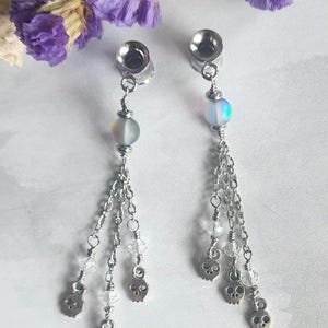 May include: A pair of silver-toned ear gauges with dangling chains. Each gauge features a clear bead, a small skull charm, and a cluster of clear crystals. The gauges are displayed against a white background with purple flowers.