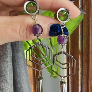 May include: A pair of silver-toned earrings with a geometric design. Each earring features a purple gemstone, a series of nested hexagonal shapes, and a conical charm. The earrings are designed for stretched ear piercings.