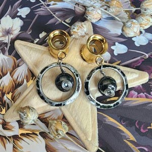 May include: A pair of gold-colored ear gauges with dangling silver-toned hoop earrings. Each earring features a dark, round bead wrapped with silver wire. The gauges and earrings rest on a light-colored, star-shaped object, against a floral background.