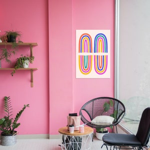 Puede incluir: Una habitación rosa con una impresión de arcoíris de colores en la pared, una silla de mimbre negra, una silla negra con un cojín blanco y una mesa de cesta de alambre blanca con dos tazas de café y una bolsa rosa.