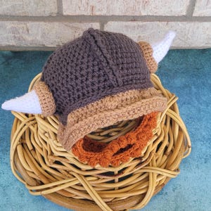 May include: A handmade crochet Viking hat in shades of gray, tan, and orange, with white horns. The hat is displayed on a round woven basket, set against a brick wall and a blue surface. This unique hat is perfect for themed costumes or photo props.
