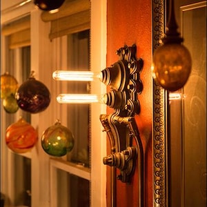 May include: A steampunk-style wall sconce with two Edison bulbs, mounted on a red wall with a gold-framed mirror behind it.
