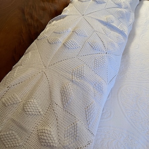 May include: White crocheted throw blanket with a diamond pattern and raised dots. The blanket is folded on a white bed.