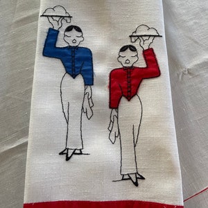 May include: A white tea towel with red trim features an embroidered design of two figures in black outlines, each carrying a tray with a cloud-like object. One figure wears a blue shirt and the other wears a red shirt.
