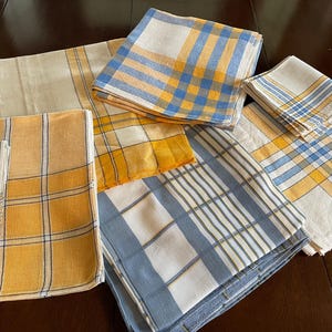May include: A collection of folded square fabric napkins featuring various plaid and checkered patterns in shades of yellow, blue, and white. The textiles are arranged in a overlapping stack, displaying different grid designs and color combinations.
