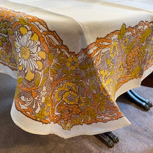 May include: A white tablecloth featuring a wide border print of brown, yellow, and olive green botanical motifs, including clusters of grapes, large flowers, and leafy vines intertwined with a dark brown cross-hatched lattice pattern.
