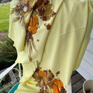 May include: A pale yellow tablecloth featuring a harvest-themed print with orange pumpkins, brown grapes, wheat stalks, and assorted nuts. The design is repeated in two corners of the fabric, showcasing a detailed autumn-inspired botanical arrangement.