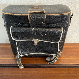 May include: A black leather camera case with a white trim and a strap. The case has a small pocket on the front.
