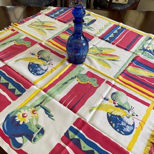 May include: A cobalt blue glass decanter with a stopper sits on a colorful tablecloth. The tablecloth features a repeating pattern of red, blue, yellow, and green geometric shapes and floral designs. The tablecloth has a fringed edge.