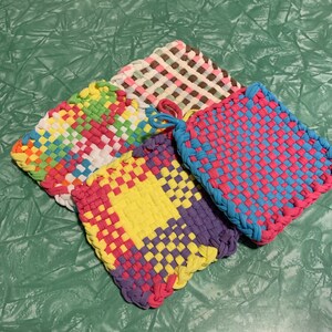 May include: A collection of colorful, woven potholders in various patterns. The potholders feature a rainbow, pink and blue, yellow and purple, and brown and white design. Each potholder has a loop for hanging.