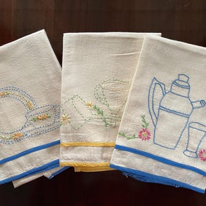 May include: Three white linen tea towels with blue and yellow trim. Each towel features a different embroidered design: a teapot and glass, a teacup and saucer, and a pitcher and glass.