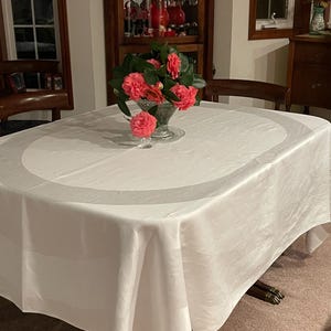 May include: A white tablecloth with a subtle circular pattern, set on a wooden table with a vase of pink camellia flowers in the center.