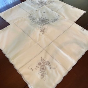 May include: Two white embroidered tablecloths with a floral design. The tablecloth has a scalloped edge and is made of a soft, delicate fabric.