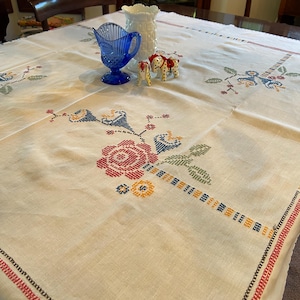 May include: A white linen tablecloth with embroidered floral designs in red, blue, yellow, and green. The tablecloth has a black and red border. There are two small white ceramic horses and a blue glass pitcher on the tablecloth.